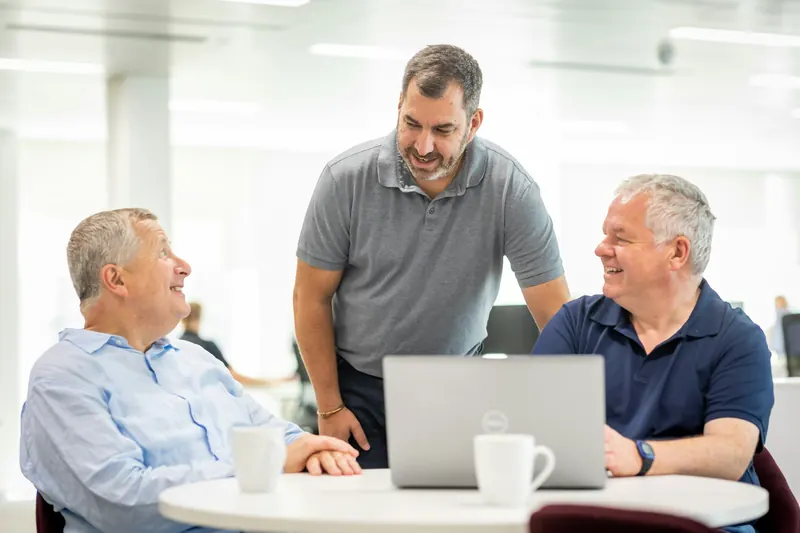 Three men talking over a laptop and mugs of tea in a bright office